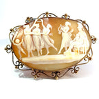 An antique-style oval carved shell or stone plaque with a gold-toned metal frame, featuring classical figures in a relief scene on a white background.