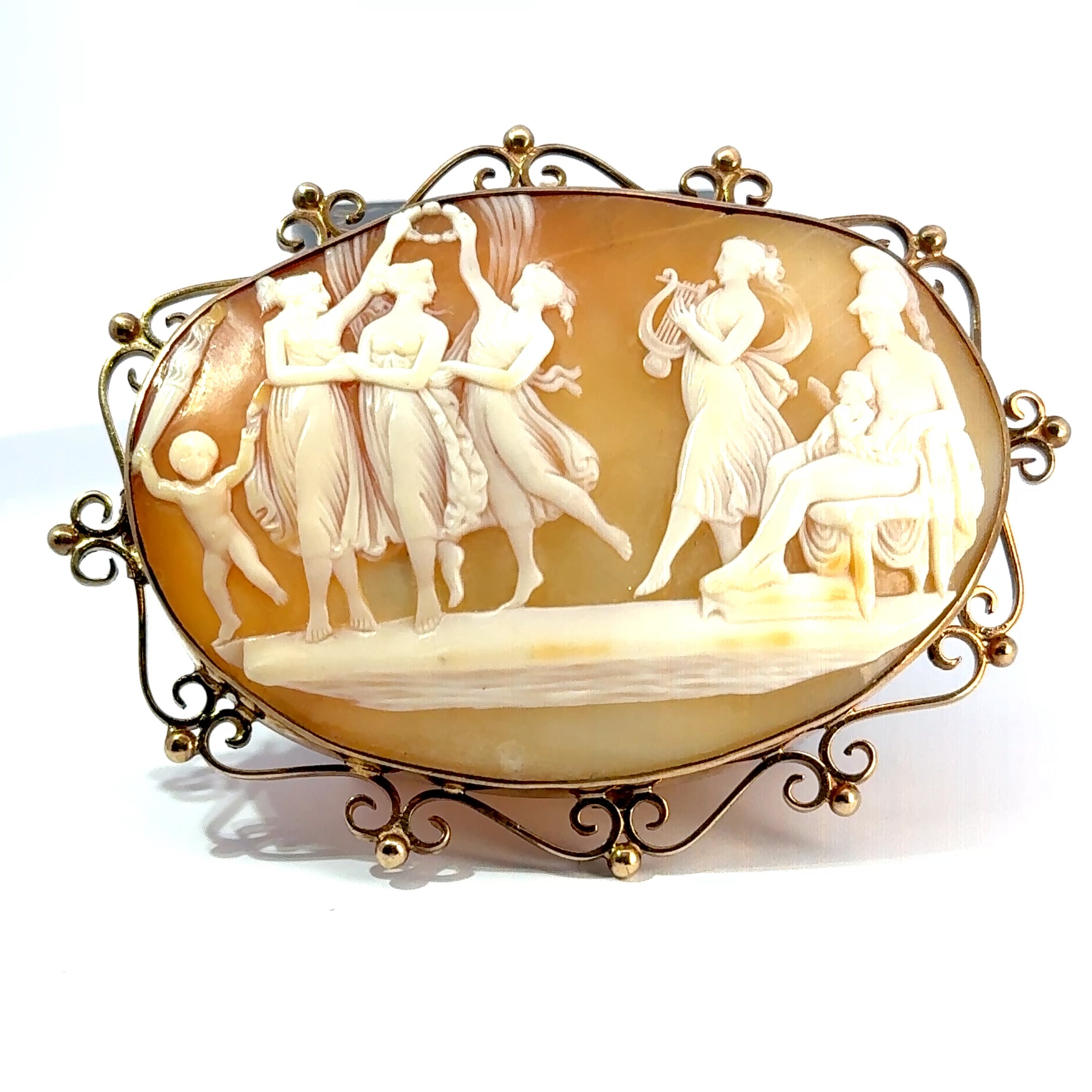 An antique-style oval carved shell or stone plaque with a gold-toned metal frame, featuring classical figures in a relief scene on a white background.
