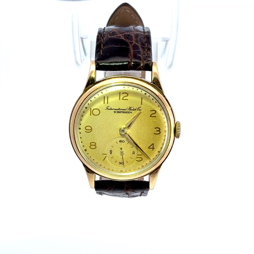 A vintage-style wristwatch with a round gold-tone case, a pale yellow dial, and a dark brown textured leather strap, shown against a plain white background.